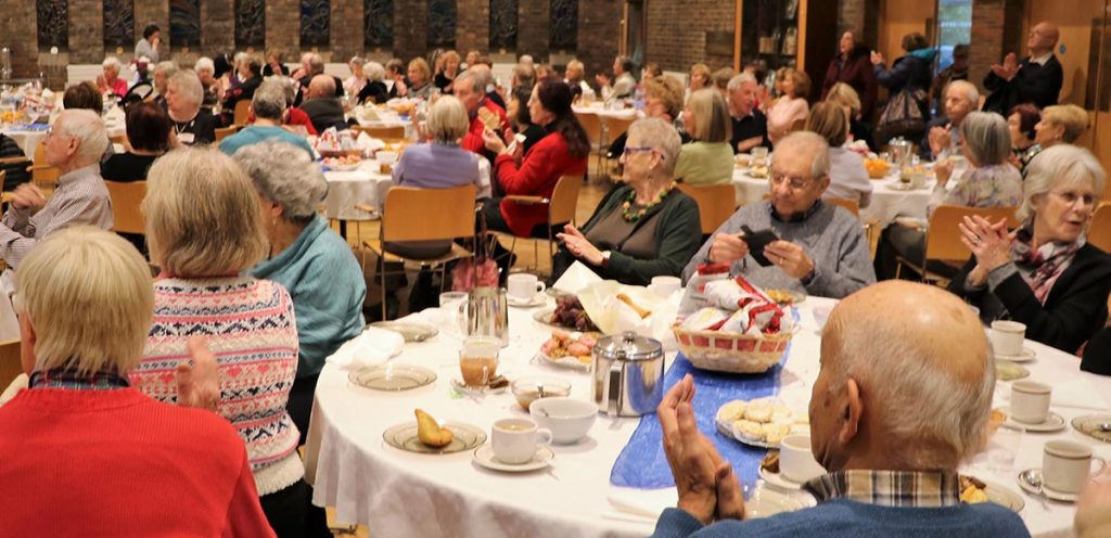 Full hall of U3A members sitting round tables laden with cakes and other food