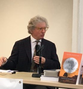 Peter Popham in a suit, sitting at a desk with a copy of his book.