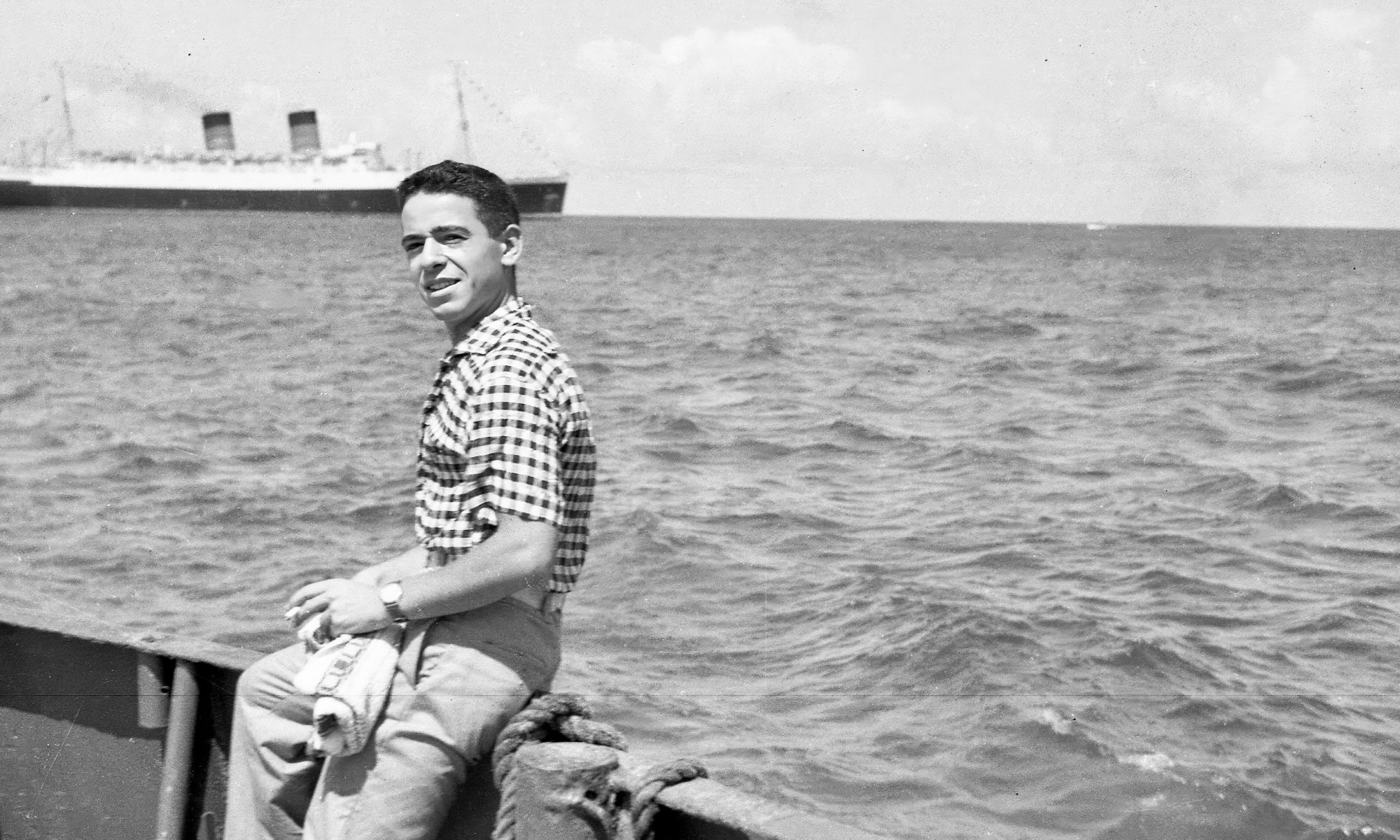Ivan Berg as a young man sitting on edge of boat with liner in background