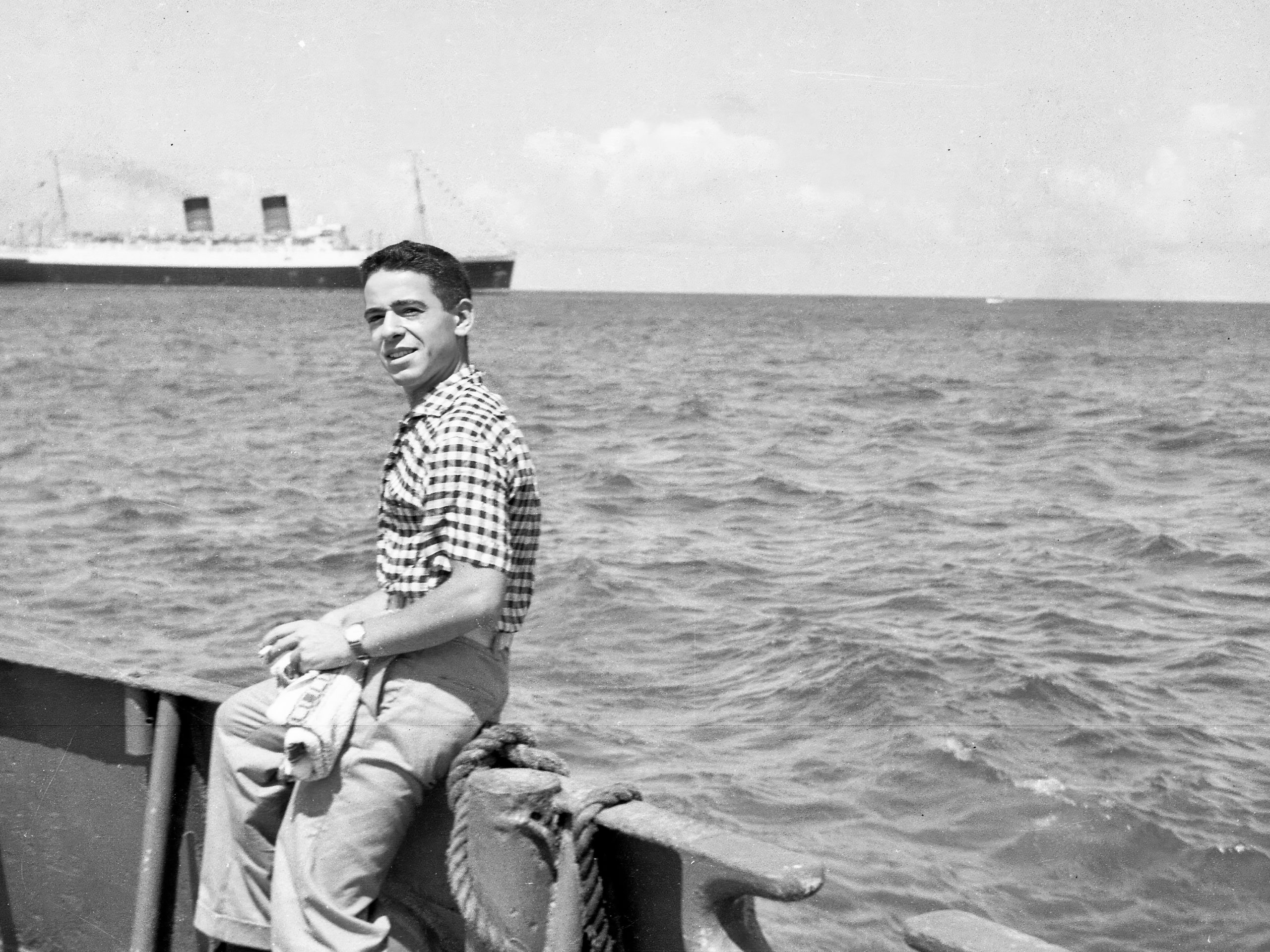 Ivan Berg as a young man sitting on edge of boat with liner in background